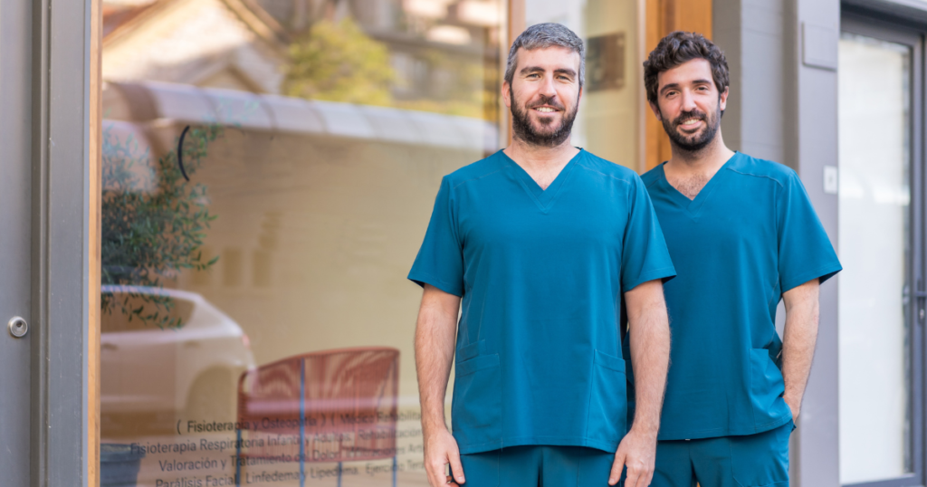 Two healthcare professionals wearing teal scrub uniforms, standing confidently outside a medical facility with glass windows.