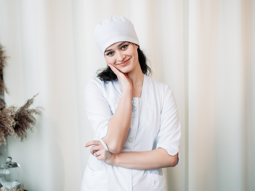 Smiling female healthcare professional wearing a crisp white scrub uniform and cap, standing in a serene environment with soft lighting.