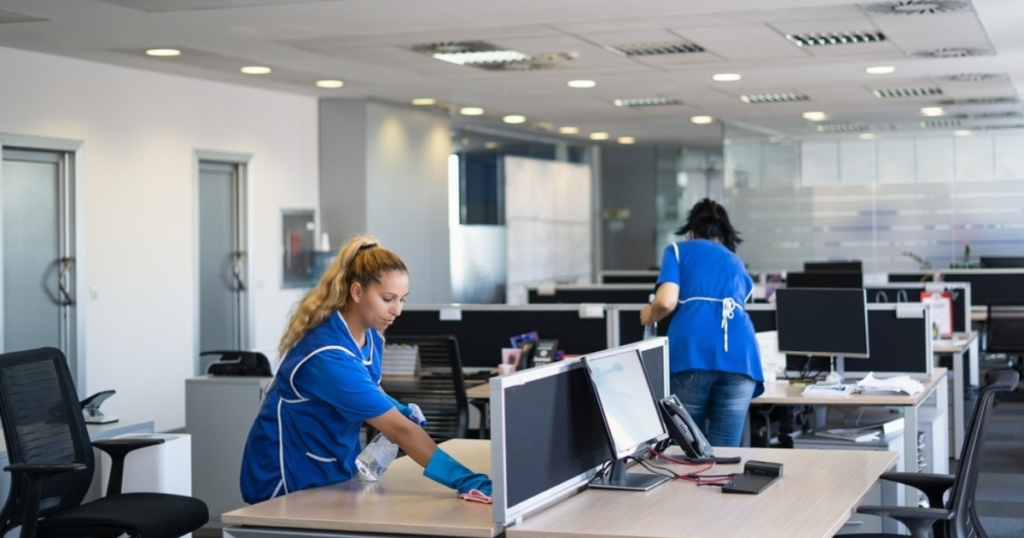 Two women wearing housekeeping cleaner uniforms are seen cleaning an office. One is wiping a desk, while the other cleans another part of the room, representing efficient and professional housekeeping services.