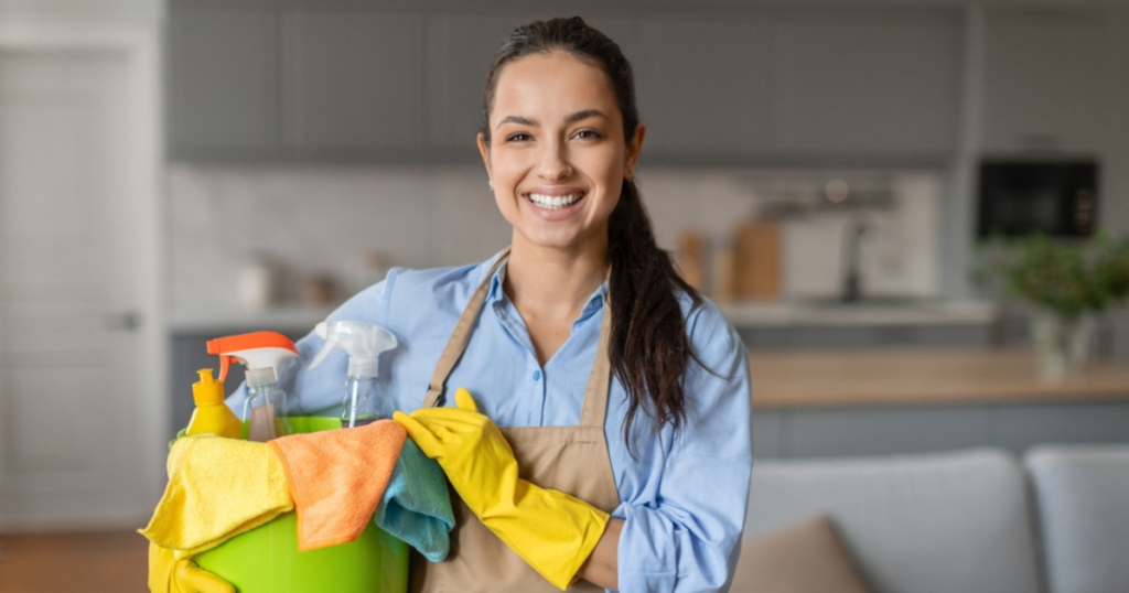 A smiling woman dressed in a housekeeping cleaner uniform holds a bucket filled with cleaning supplies. She is wearing gloves and an apron, reflecting professionalism in her role as a cleaner.