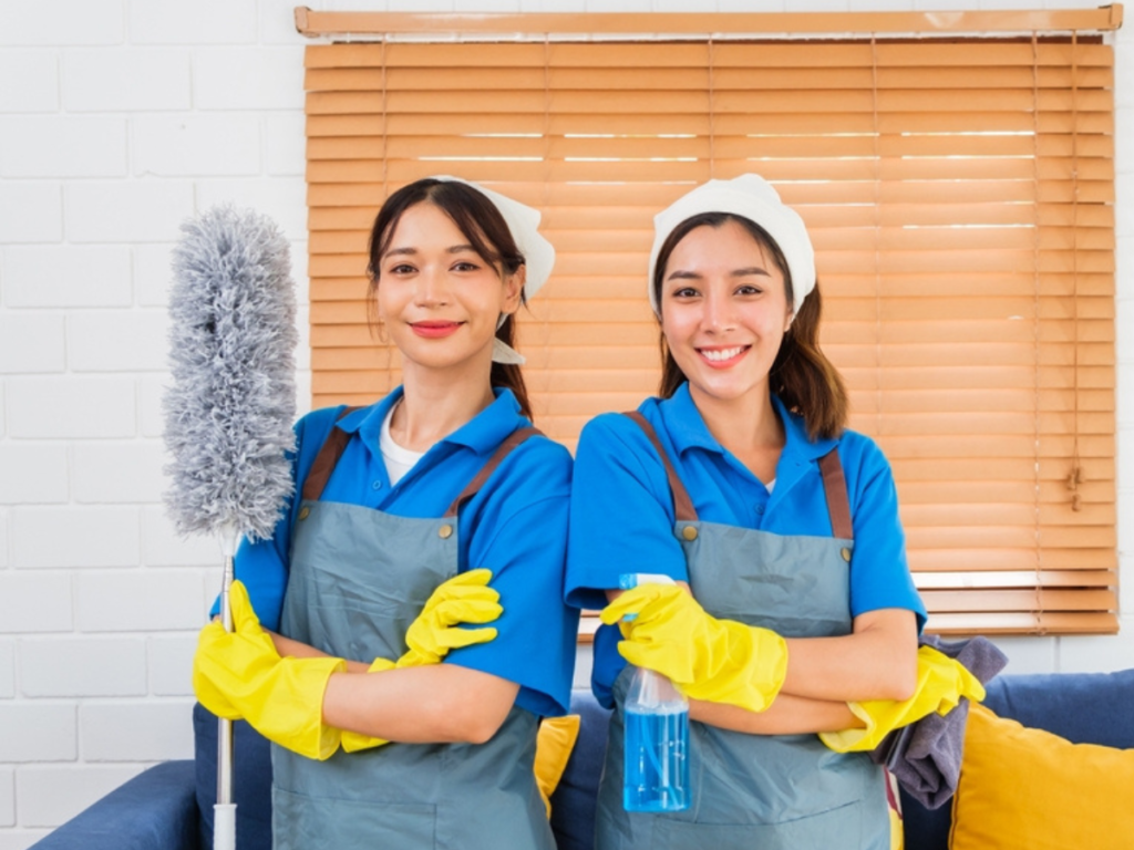 Two women wearing housekeeping cleaner uniforms, including blue shirts, gray aprons, and yellow rubber gloves, standing with cleaning tools. One woman holds a duster, and the other holds a spray bottle and cloth. They are smiling and standing in front of a window with wooden blinds.