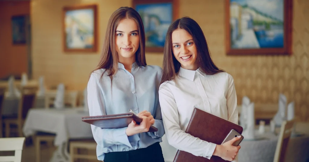 Two female staff members in a restaurant environment wearing professional staff uniforms while holding clipboards.