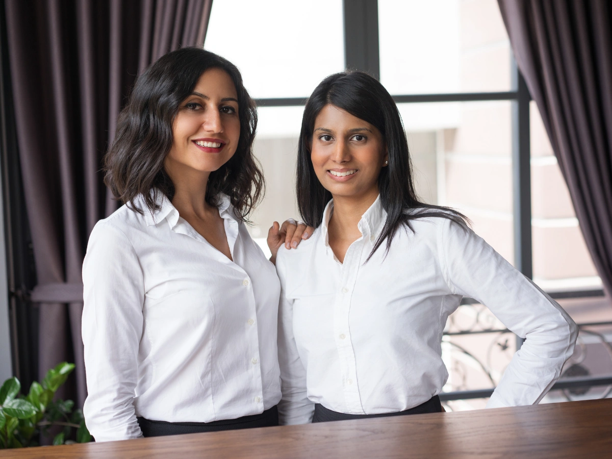 Two female hospitality staff members standing in formal attire, wearing coordinated white staff uniforms.