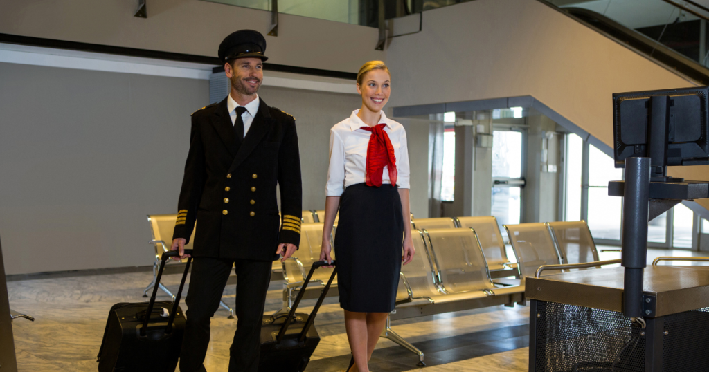 Smiling pilot and flight attendant in professional uniforms walking through an airport terminal with luggage.